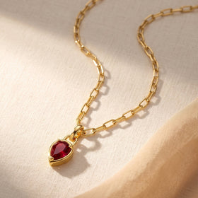 A yellow gold link necklace with a red ruby pendant lies diagonally across cream woven fabric.