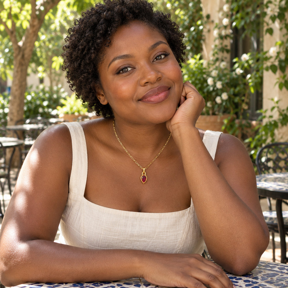 A woman with short curly hair wears a gold link necklace with a red pear-shaped pendant at a leafy outdoor cafe.