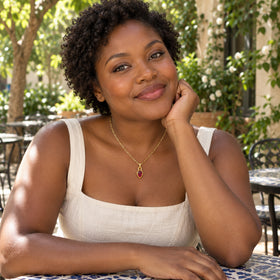 A woman with short curly hair wears a gold link necklace with a red pear-shaped pendant at a leafy outdoor cafe.