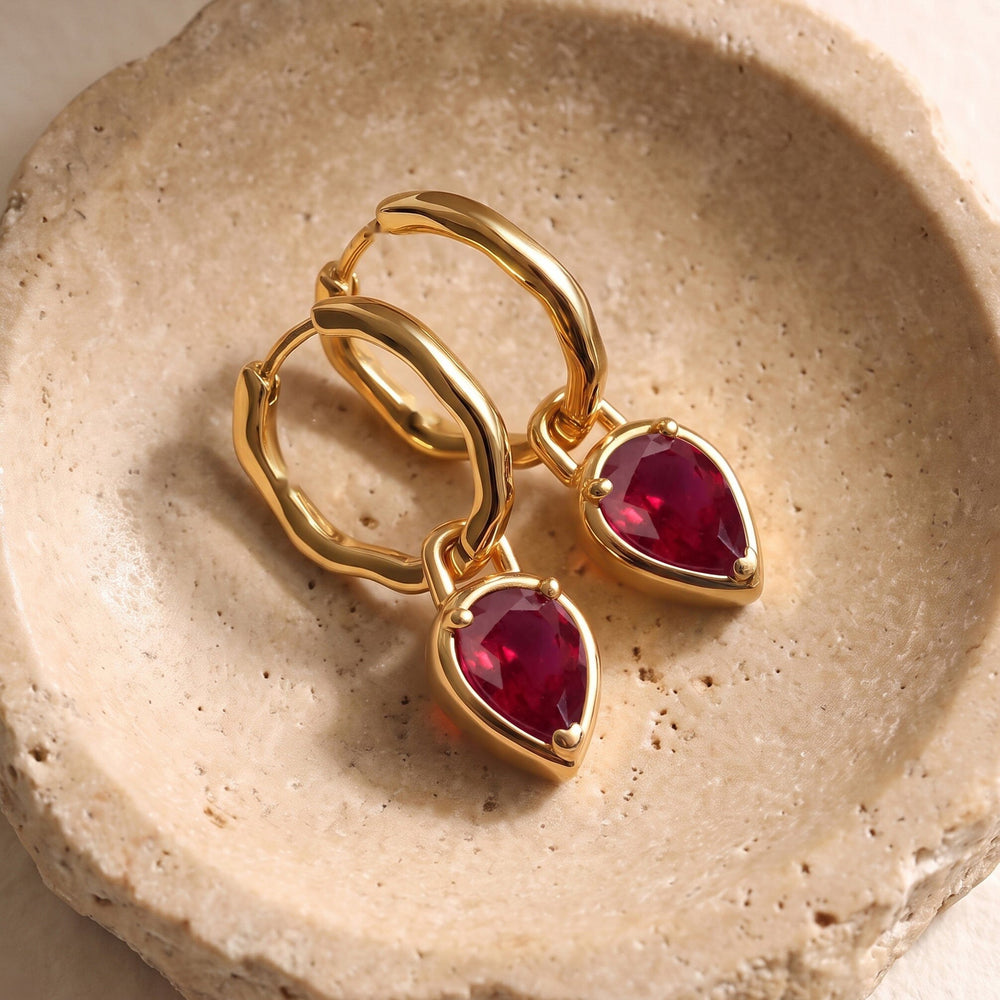 Yellow gold plated ruby hoops rest diagonally together on a pale textured stone dish.