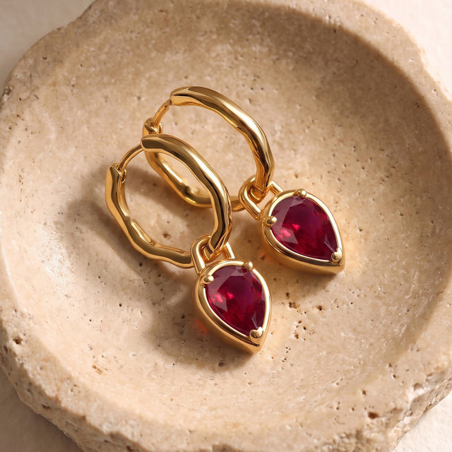 Yellow gold plated ruby hoops rest diagonally together on a pale textured stone dish.