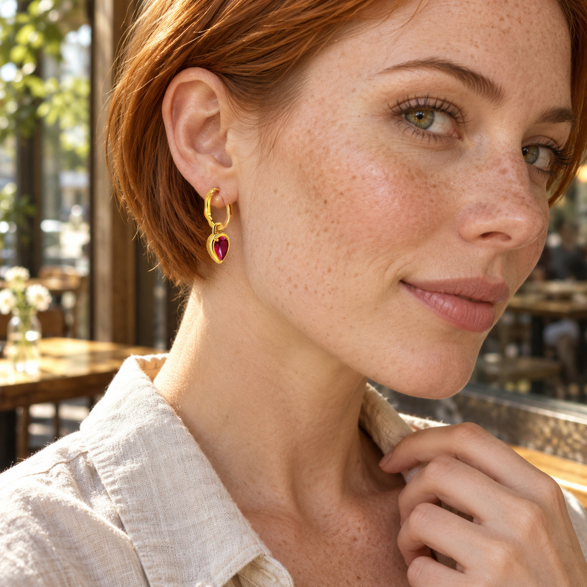 A woman with copper hair wears a yellow gold plated ruby hoop beside a sunlit cafe window.