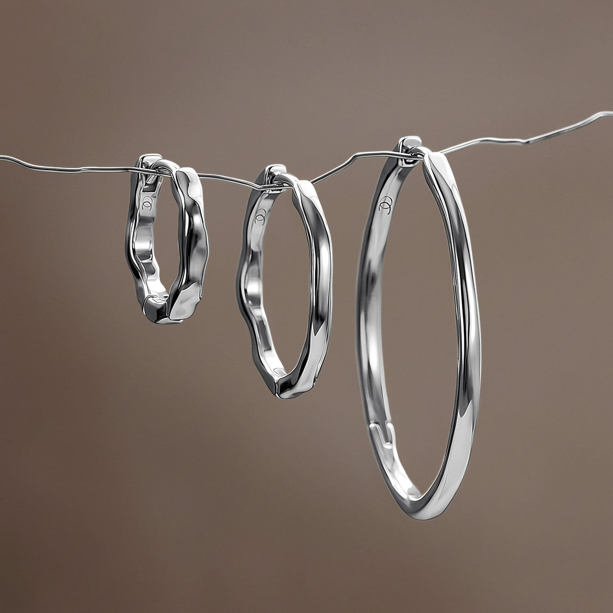 Three graduated wavy hoop earrings hang from a thin wire against a softly blurred brown background.