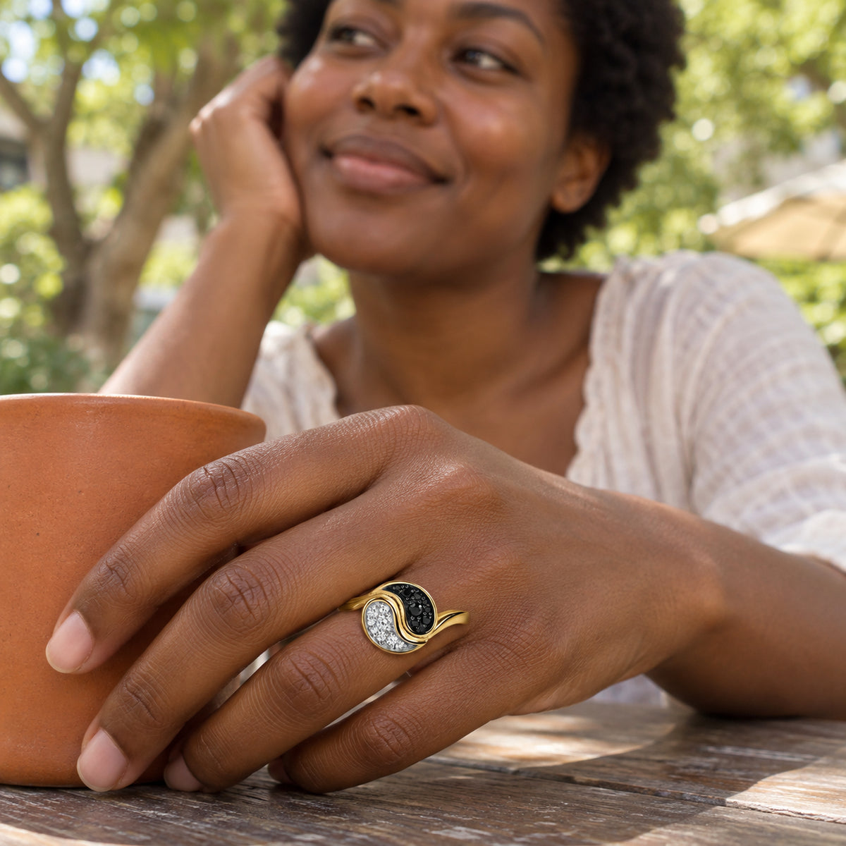 A yellow gold plated ring is worn on a hand beside a terracotta cup at an outdoor cafe.