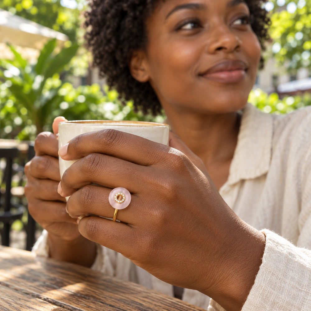 A yellow gold vermeil ring with a pink rose quartz disc is worn on a hand holding a ceramic cup at a leafy outdoor cafe.