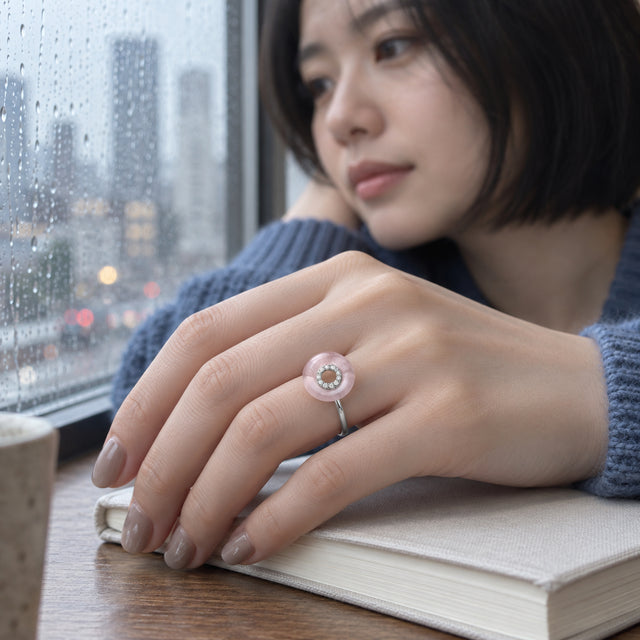 A sterling silver ring with a pale pink disc and clear stones is worn on a hand resting on a notebook beside a rain-covered window.