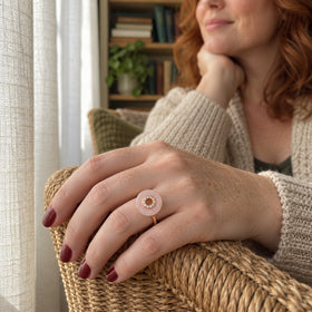 A rose gold vermeil ring with a circular pink stone is worn on a freckled hand resting on a woven chair beside a linen curtain.