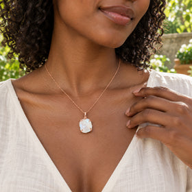 A rose gold moonstone pendant hangs from a fine chain above an ivory blouse in a leafy garden.