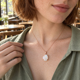 A moonstone pendant with rose gold prongs rests on a freckled neckline above a sage blouse in a cafe.