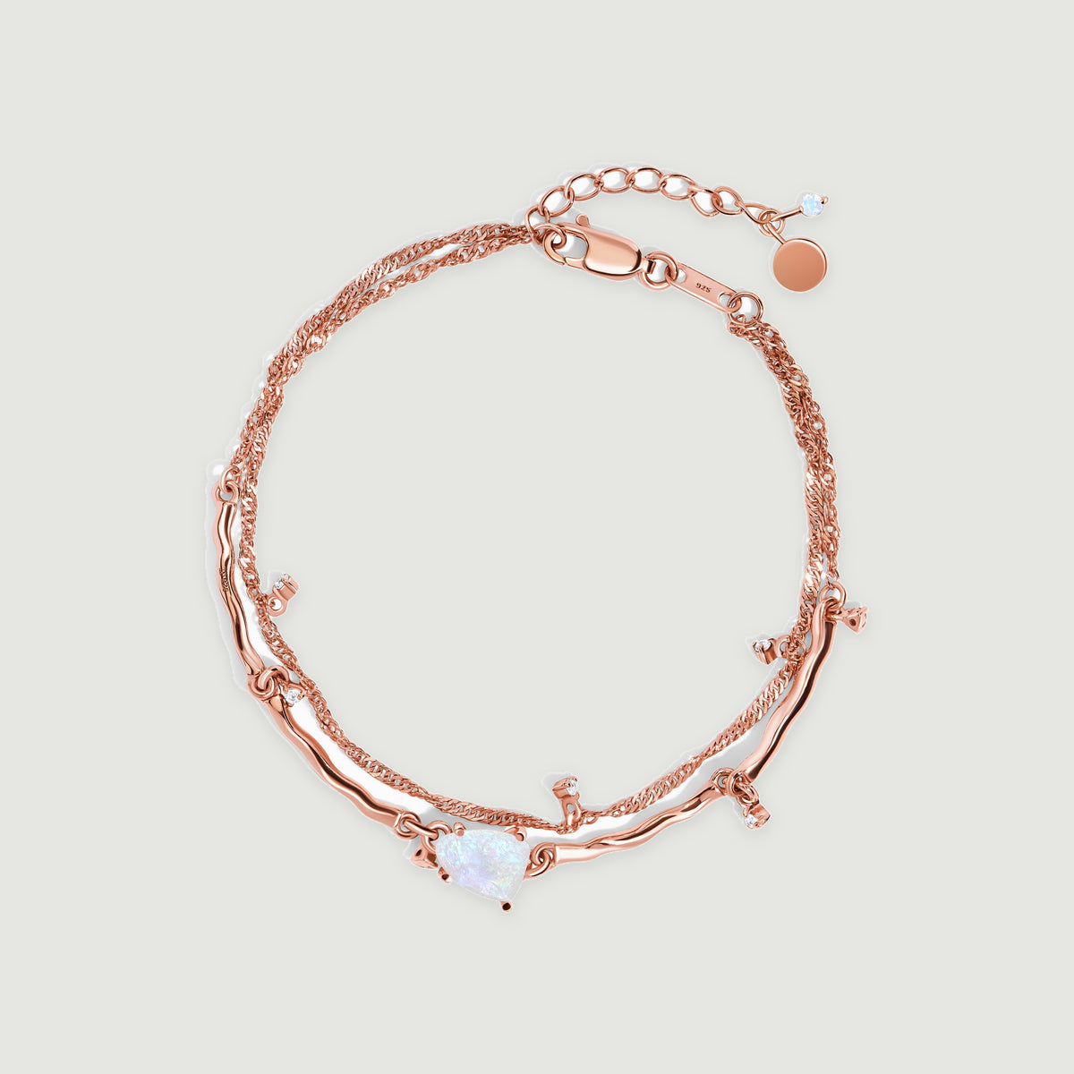 A rose gold vermeil bracelet lies in an oval on light gray, showing its pale moonstone, fine double chain and small clear accents.