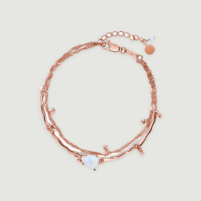 A rose gold vermeil bracelet lies in an oval on light gray, showing its pale moonstone, fine double chain and small clear accents.