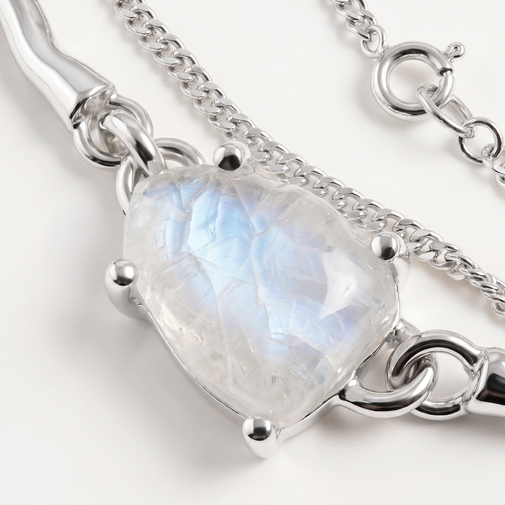 A close view shows blue reflections inside the pale moonstone beside silver prongs, a polished link and a fine chain on a clean tabletop.