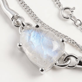 A close view shows blue reflections inside the pale moonstone beside silver prongs, a polished link and a fine chain on a clean tabletop.
