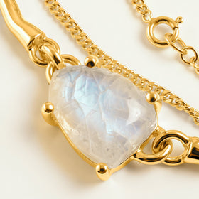 Yellow gold prongs surround an irregular milky moonstone with a blue sheen, with polished links and a fine chain visible on the tabletop.