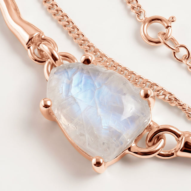 An irregular translucent moonstone catches a soft blue reflection between rose gold prongs beside the bracelet chain on a clean tabletop.