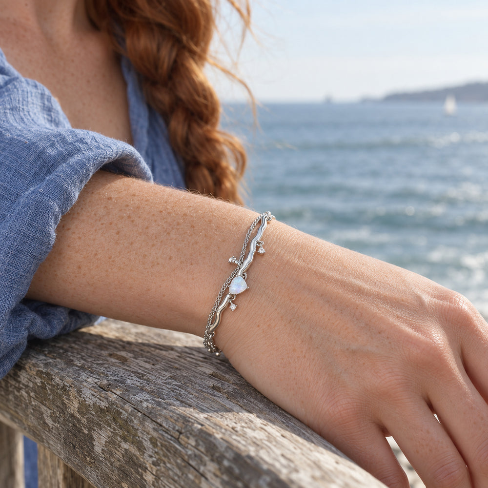 A silver moonstone bracelet with wavy links and a fine chain is worn on a freckled wrist resting on a wooden seaside railing.
