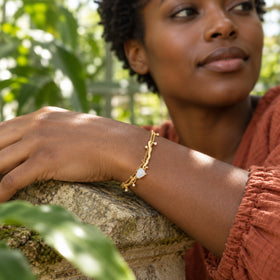 A yellow gold moonstone bracelet is shown on a dark-skinned wrist resting on a stone wall among green leaves.