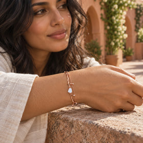 A rose gold moonstone bracelet is worn on a tan wrist resting on a stone ledge in a sunlit courtyard.