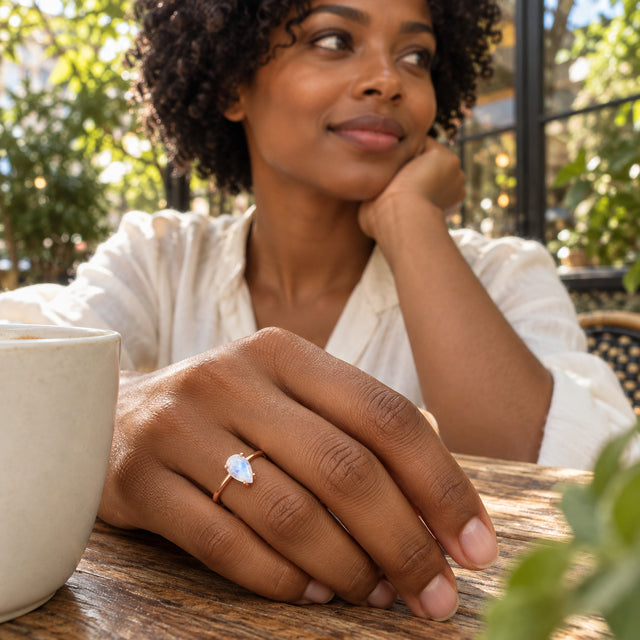 A woman with curly hair wears a pear-shaped moonstone ring on her hand beside a ceramic cup at an outdoor cafe.