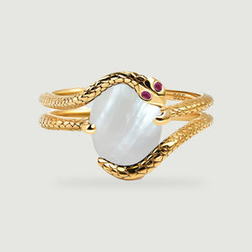 A yellow gold wrap ring with an oval white center and two small red stones rests on a light gray background.