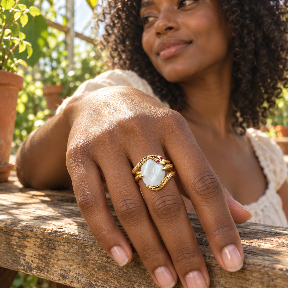 A yellow gold wrap ring with a pearly white center is worn on a hand resting on a wooden bench among green plants.